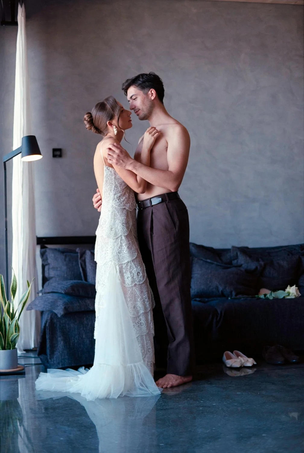 A couple sharing an intimate moment in a bedroom. The woman is wearing a lace dress and the man is shirtless, with his trousers pulled down slightly. They are standing close, foreheads touching, with soft lighting and a dark bed in the background.