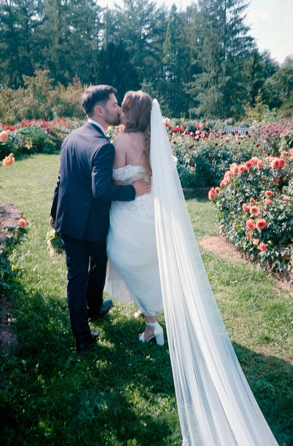 A bride and groom kissing in a garden with blooming flowers and trees in the background.