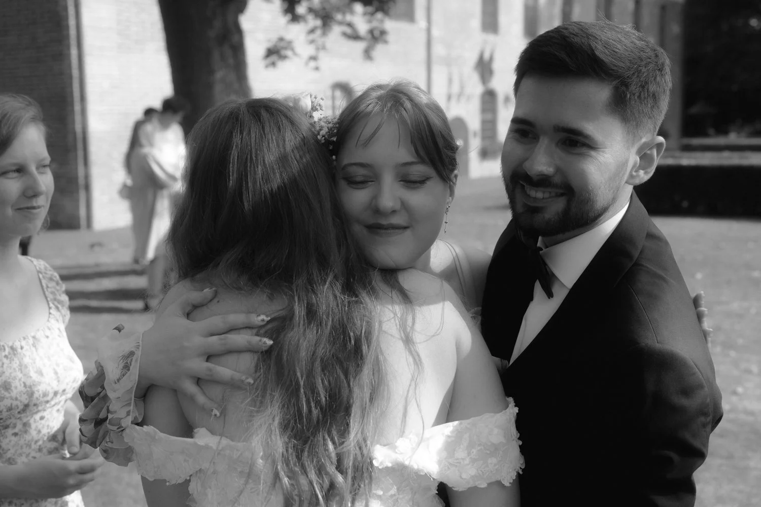 A group of people at a wedding, with a woman in a wedding dress hugging a man in a tuxedo, both smiling, while others look on happily.
