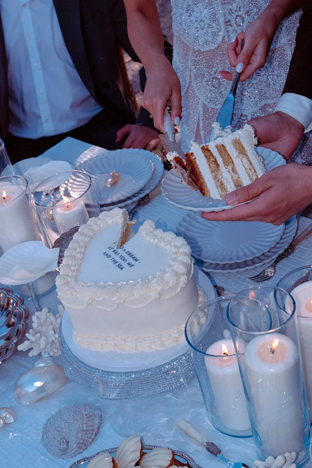 People cutting a slice of a layered wedding cake with white frosting, surrounded by candles and decorated table.