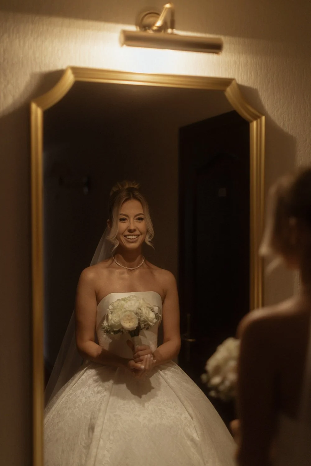 A woman in a wedding dress holding a bouquet, smiling as she looks at herself in a mirror.