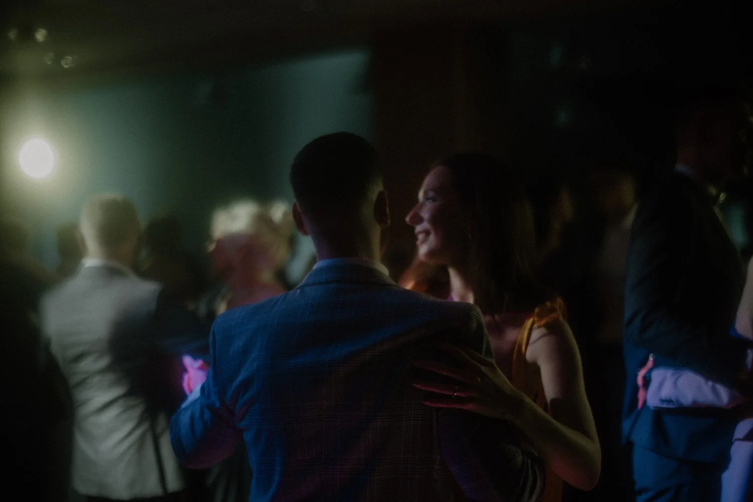 Couple dancing at a party or event in a dimly lit room with other people dancing and socializing.