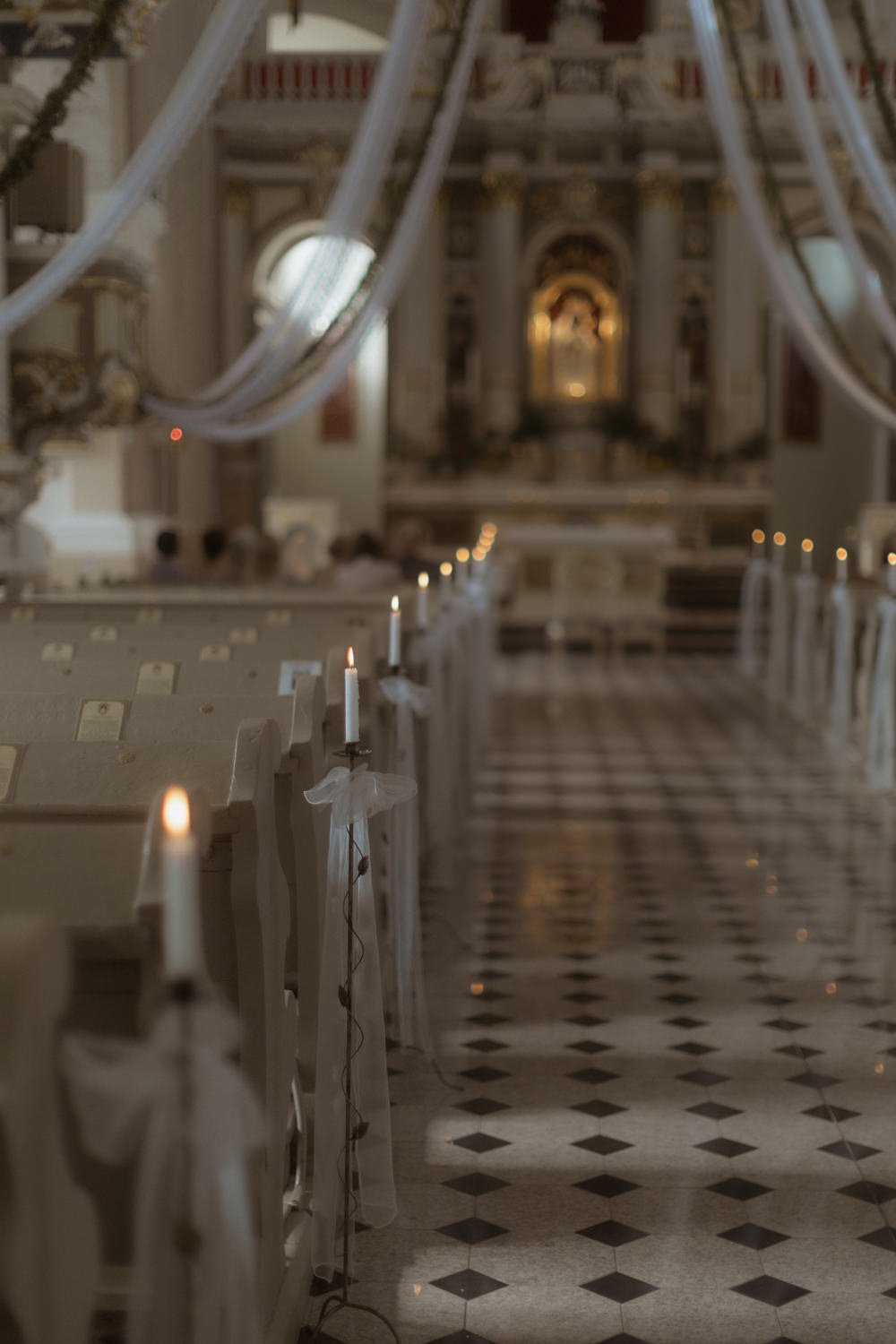Interior of a church decorated with candles attached to the pews with white ribbons, leading to an ornate altar in the background, illuminated by soft lighting.