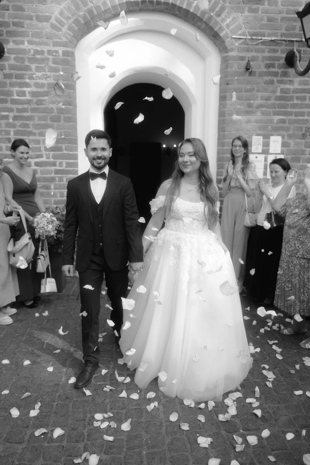 A newlywed couple walking out of a church, holding hands, as guests throw flower petals. The bride is in a white wedding dress, and the groom is in a black tuxedo. Guests are standing on either side, applauding and smiling.