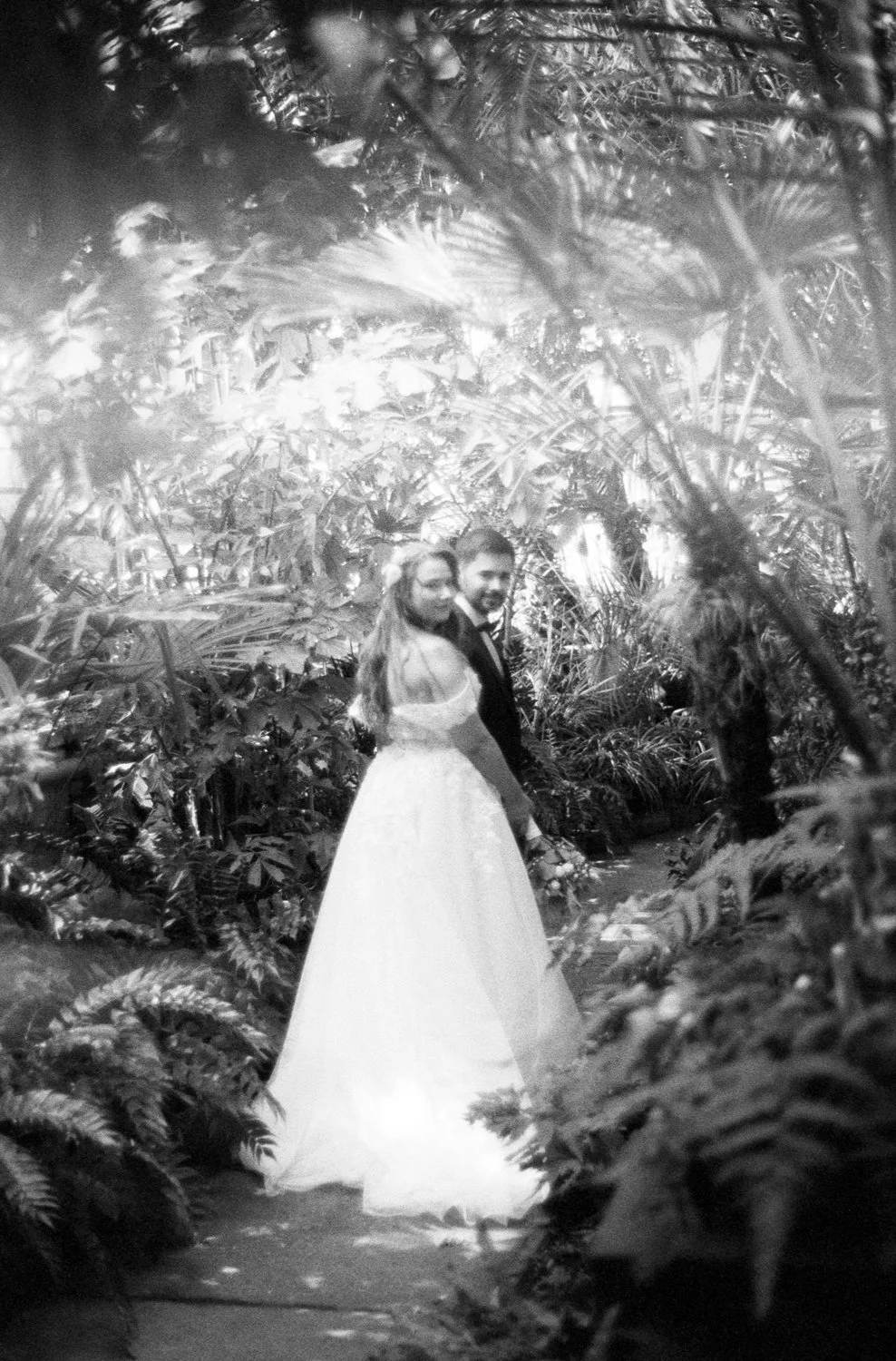 A black and white photo of a bride and groom standing together in a lush, jungle-like setting. The bride is wearing a long wedding dress, and the groom is in a suit. They are looking back over their shoulders at the camera, surrounded by dense tropic