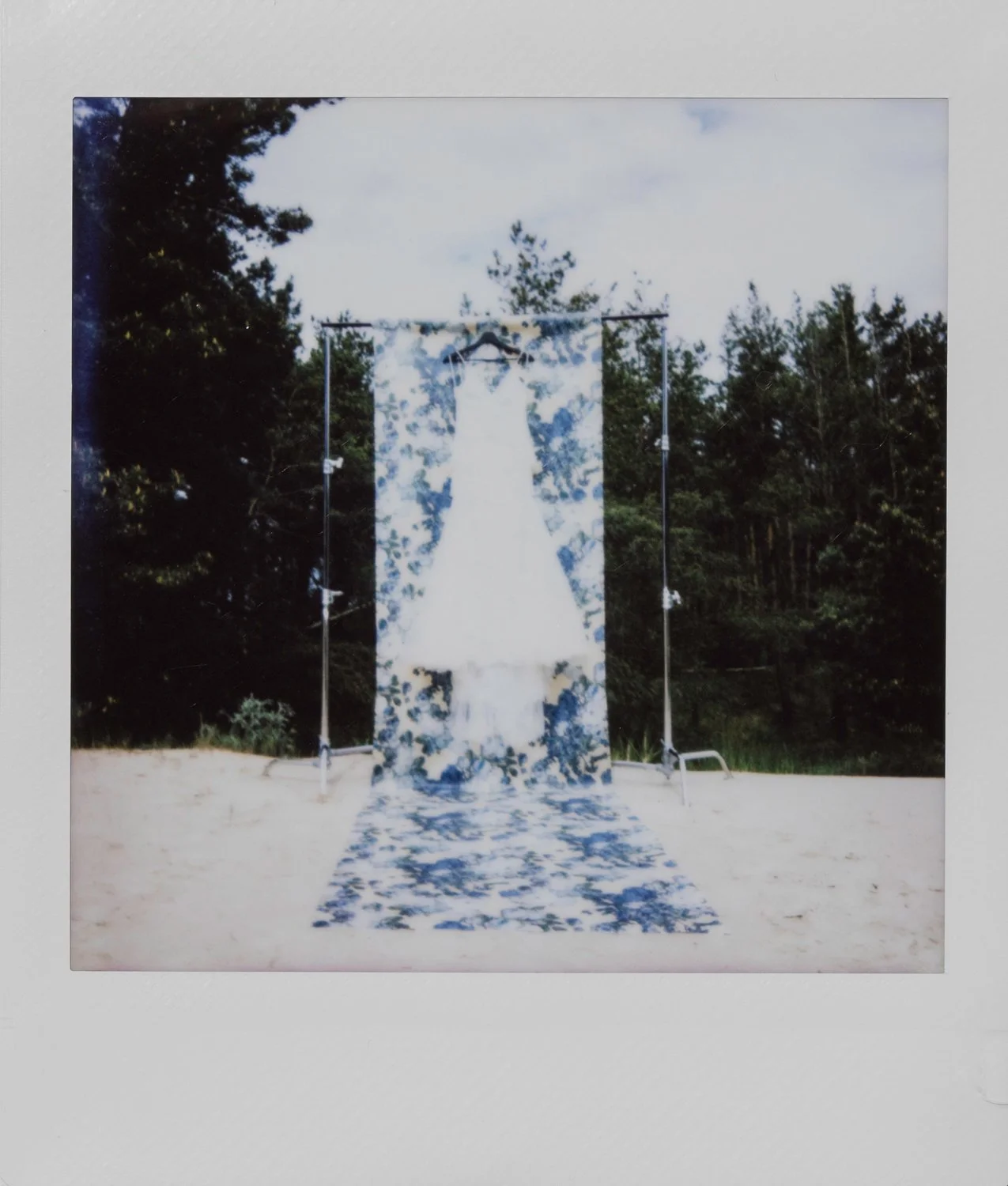 Wedding dress hanging on a hanger in front of a blue and white floral backdrop with matching runner, outdoors on a sandy surface with trees in the background.