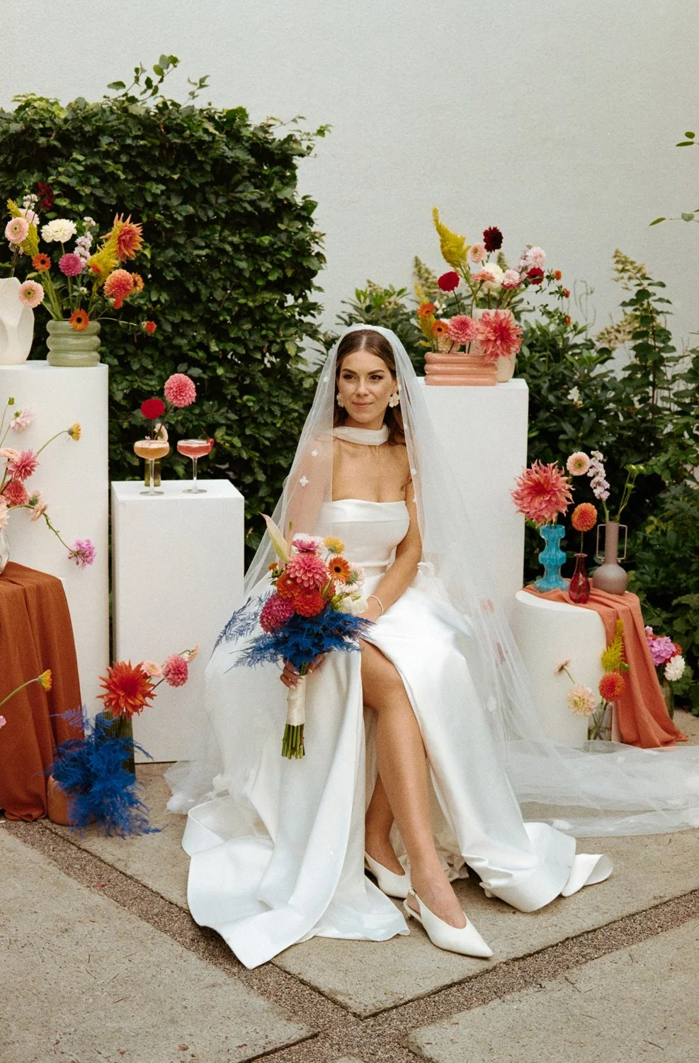 A bride sitting on a white chair at her wedding, holding a vibrant bouquet of pink, red, and blue flowers. She is wearing a white wedding dress with a veil, surrounded by colorful flower arrangements and vases on various pedestals.