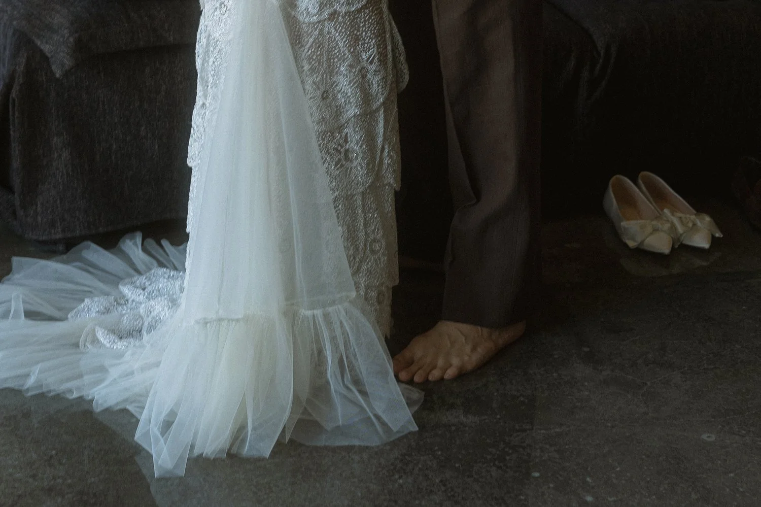 Part of a bride's wedding dress, an older person's feet, and wedding shoes on the floor.
