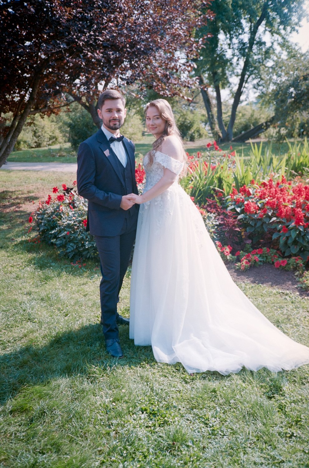 A bride and groom holding hands standing on grass in a garden with colorful flowers and trees in the background.