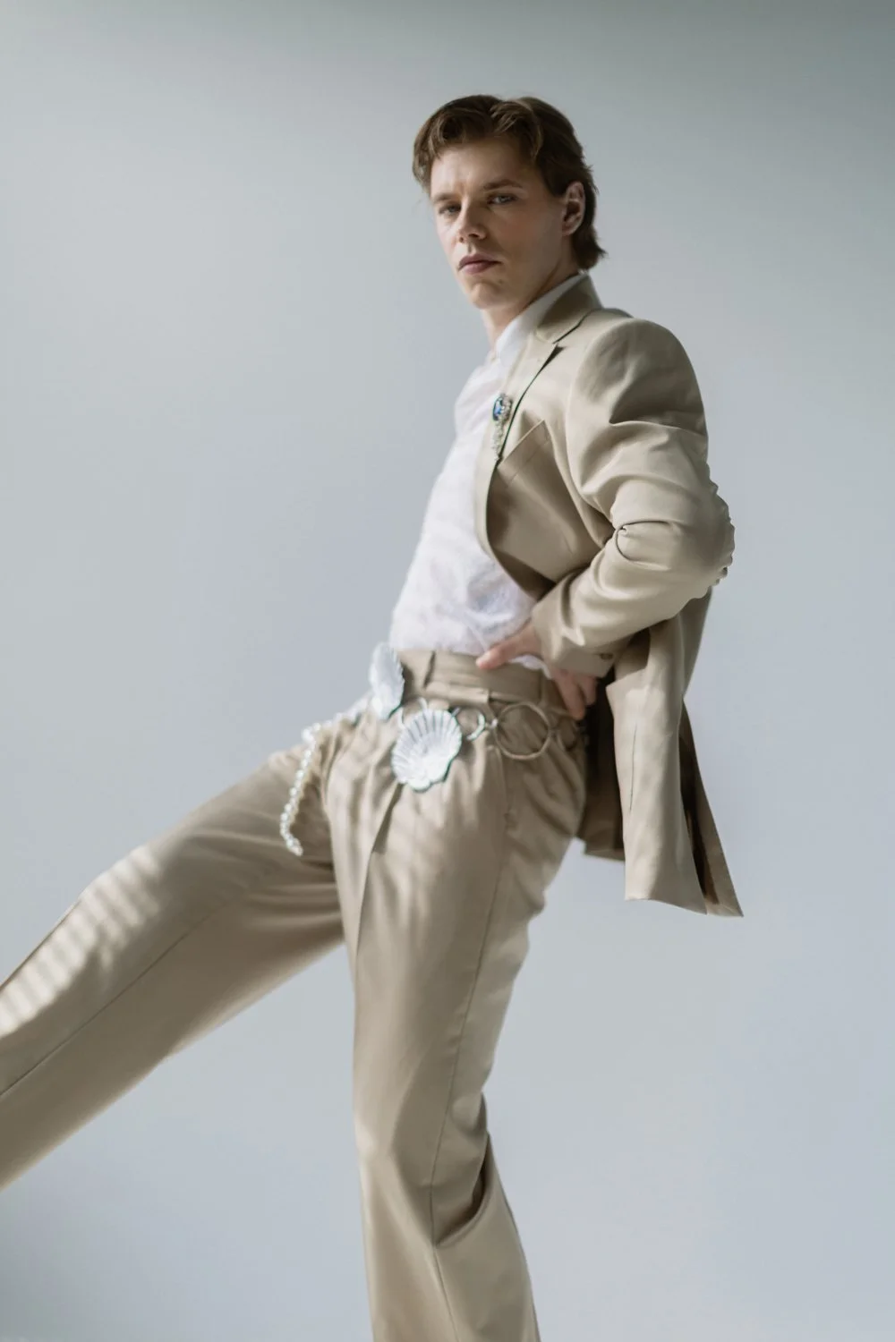 A young man with brown hair, wearing a beige suit jacket and matching pants, poses with one hand on his waist against a plain gray background.