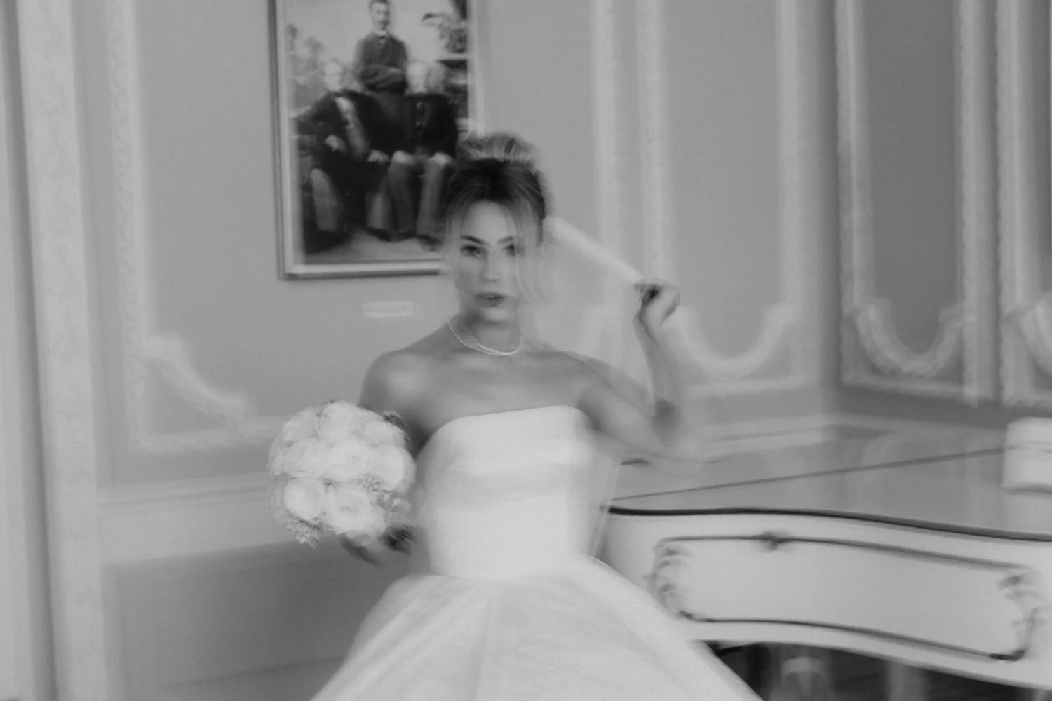 Black and white photo of a woman in a wedding dress holding a bouquet, standing in a decorated room with curtains and a framed painting on the wall.
