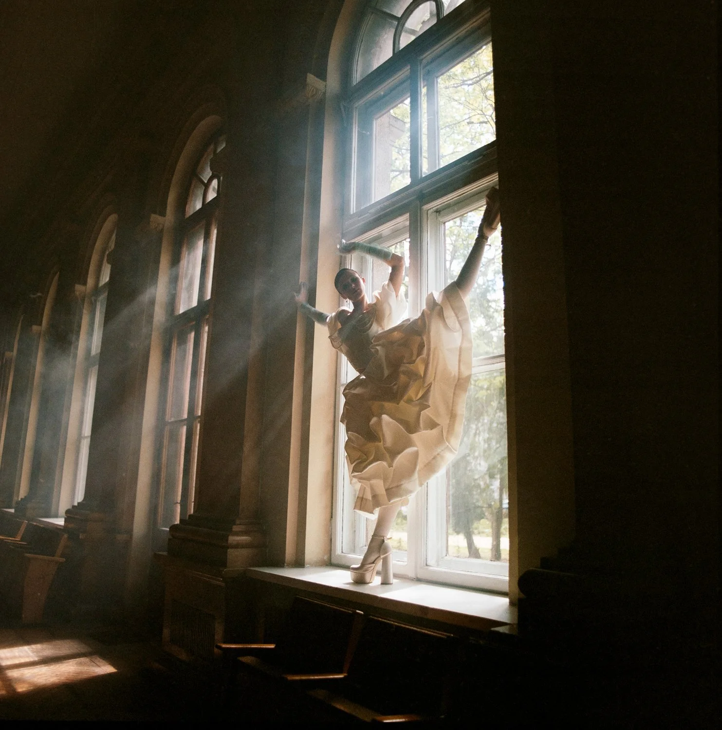 A woman in a flowing dress and high heels standing on a window sill with sunlight streaming through large windows, casting shadows in a dimly lit room.