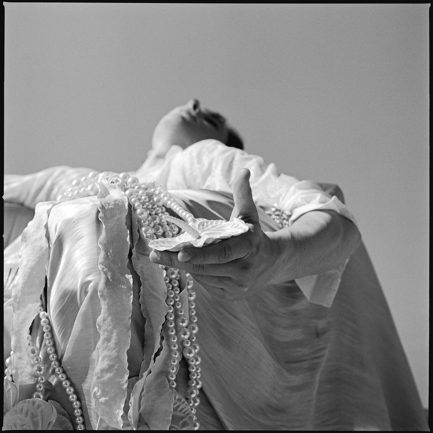 A person holding a small dish with a straw, surrounded by pearl necklaces and ribbons, with their face turned and partially out of the frame.