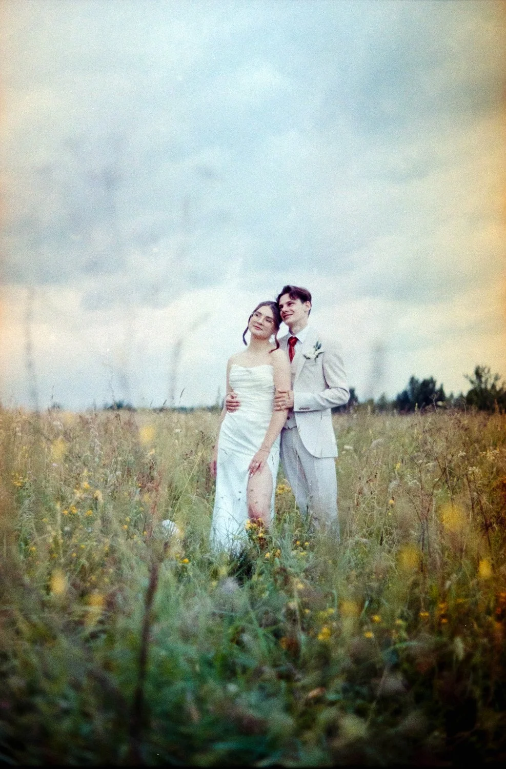 A bride and groom standing in a grassy field, smiling, with the bride in a strapless white wedding dress and the groom in a light-colored suit with a red tie, under a cloudy sky.