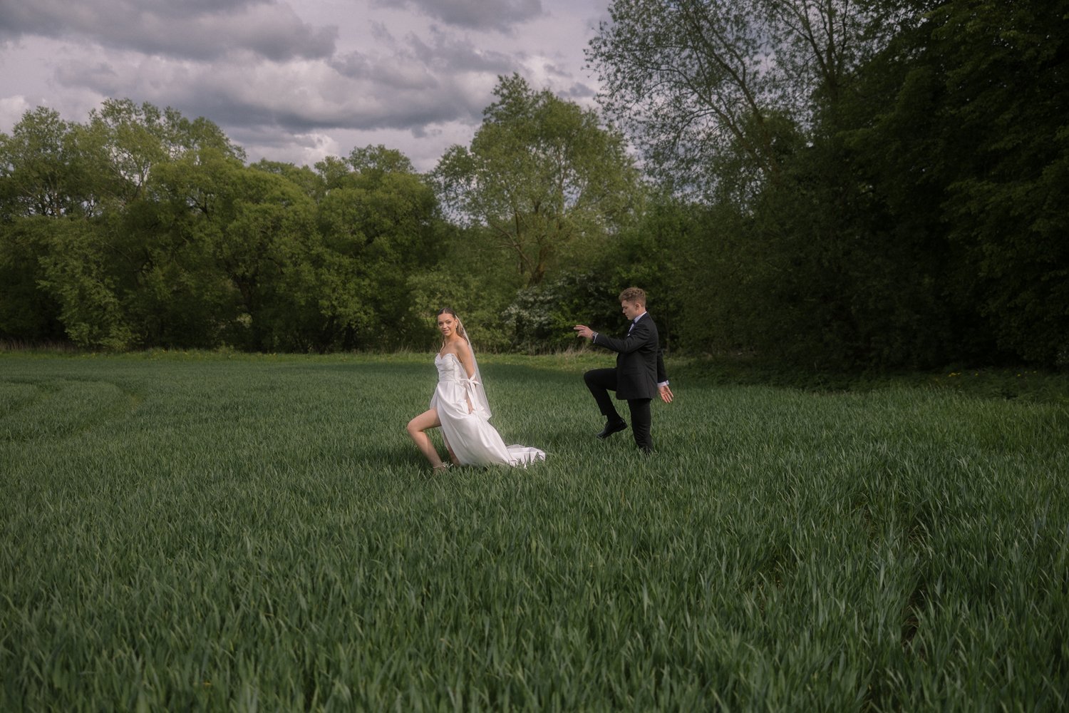 A bride in a white wedding dress and a groom in a black suit pose on a grassy field with trees and a cloudy sky in the background.