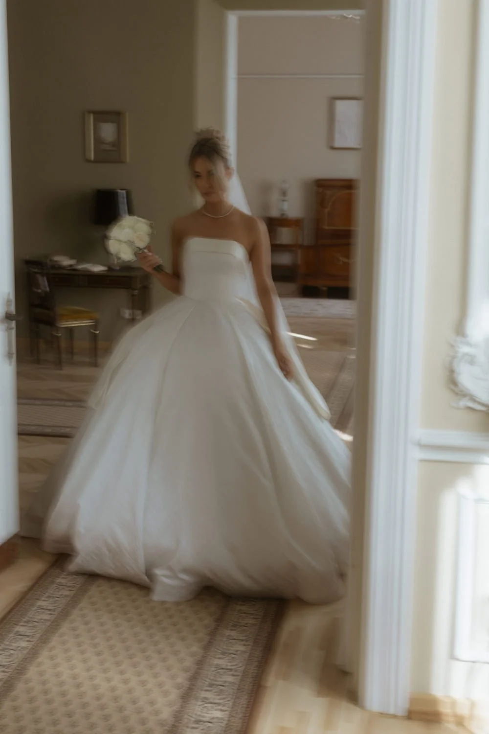 A woman in a white wedding dress holding a bouquet of white flowers, standing in front of a mirror taking a selfie indoors.