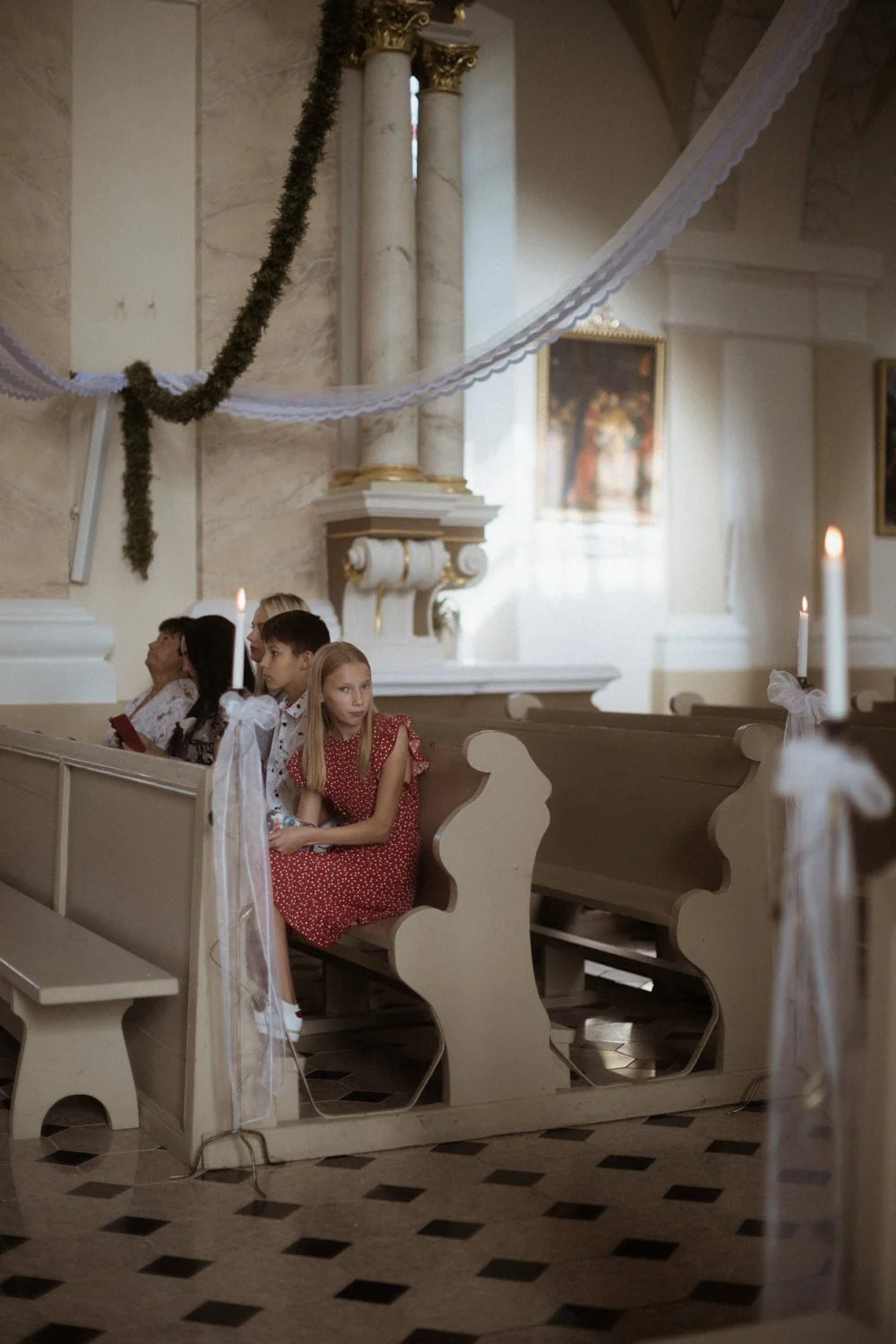 Children sitting in church pews decorated with candles and white ribbons, with church interior decor visible in the background.