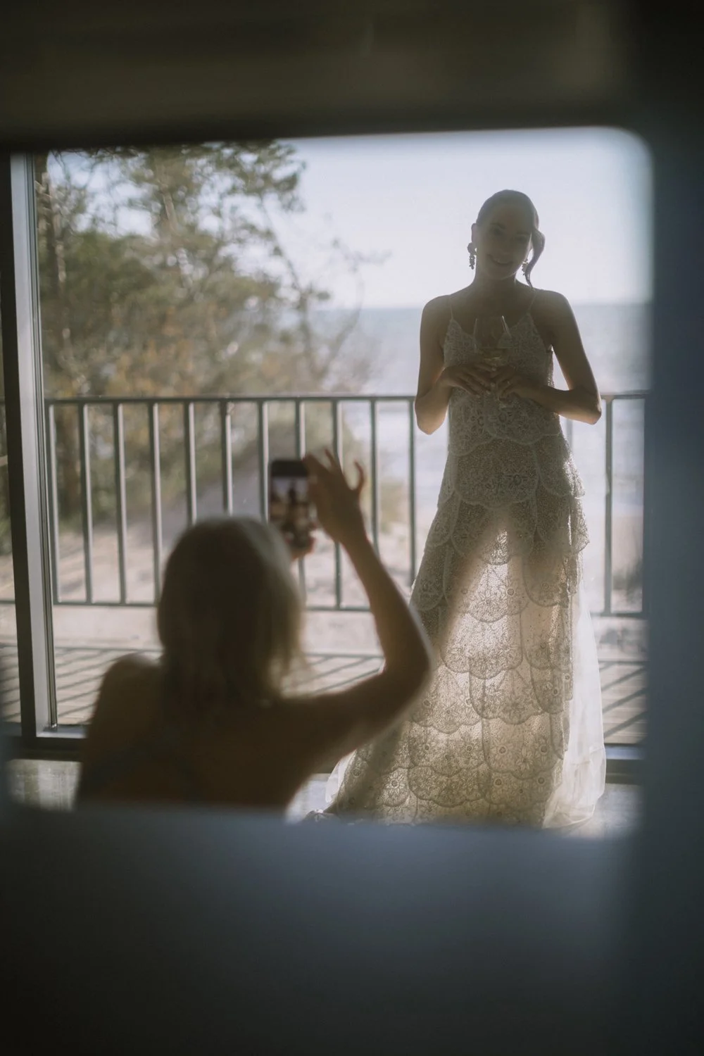 A woman in a lace dress holding a wine glass, smiling in front of a coastal background, as another person takes a photo from inside a room.