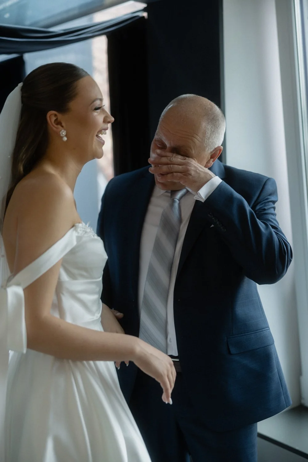 A bride in a white dress is laughing while an older man, in a suit, is covering his face and crying, standing near a window.