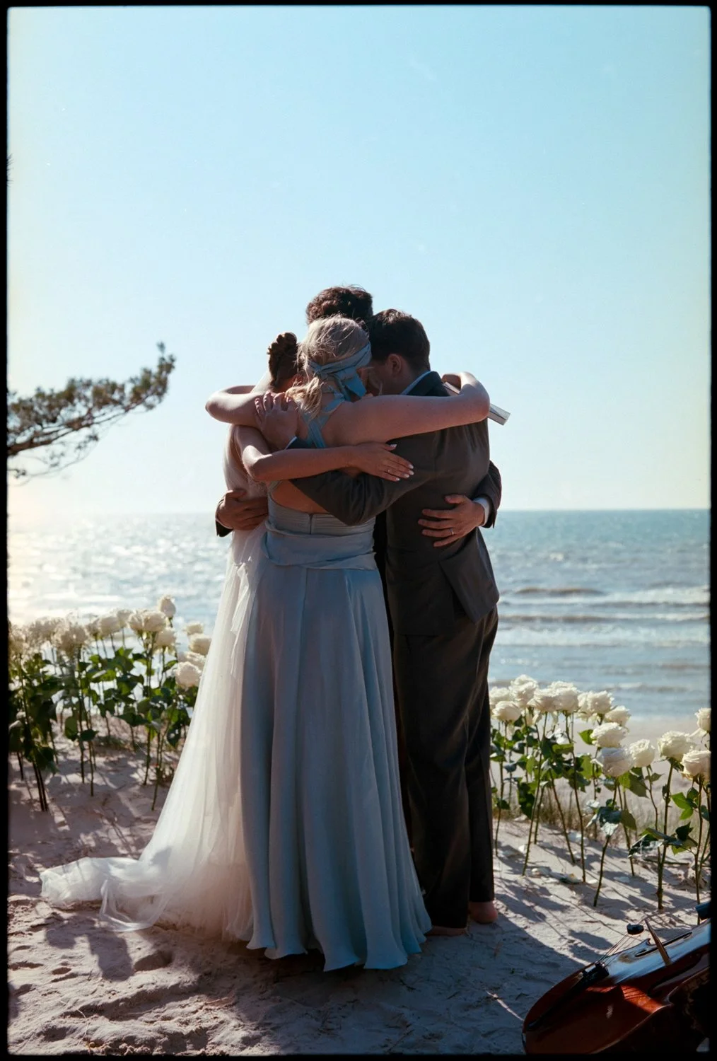 A group of people, likely a family, at elopement,  embracing each other on a beach with white flowers and the ocean in the background during sunset.