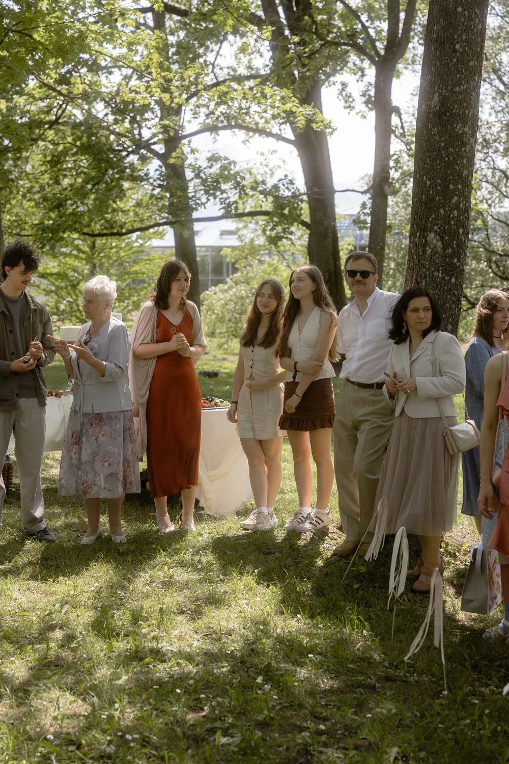 People gathered outdoors in a park on a sunny day, standing near trees, dressed in casual and semi-formal attire. A table with items is visible in the background.