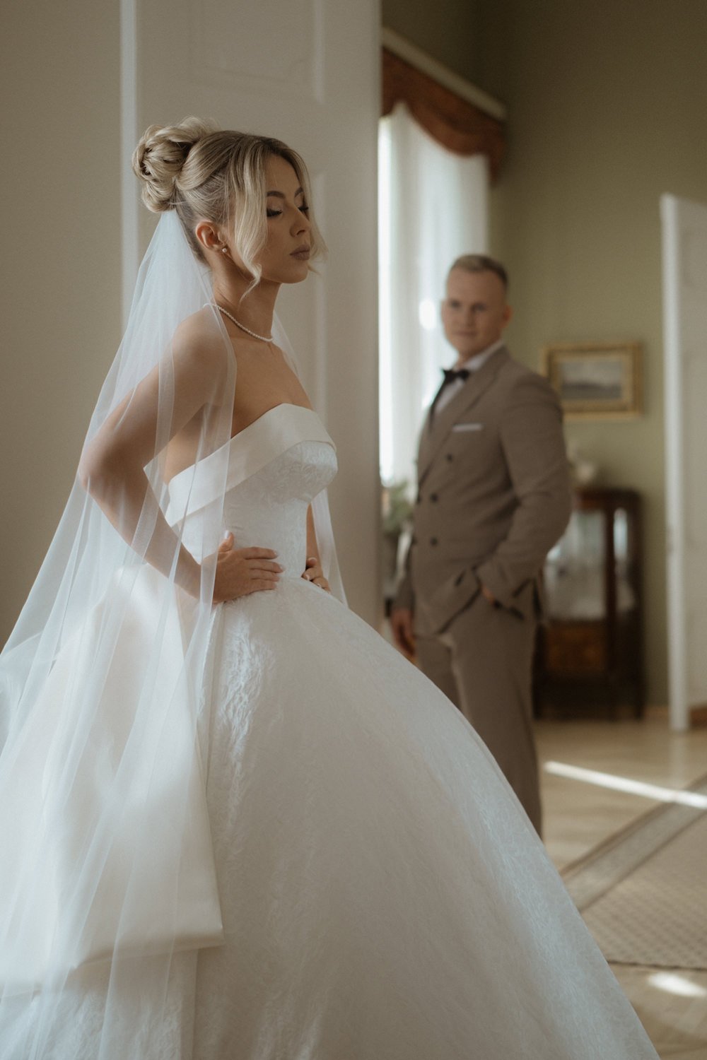 Bride in a white wedding dress with a veil, standing with her hands on her hips, and a groom in a beige suit with a bowtie in the background, inside a decorated room.