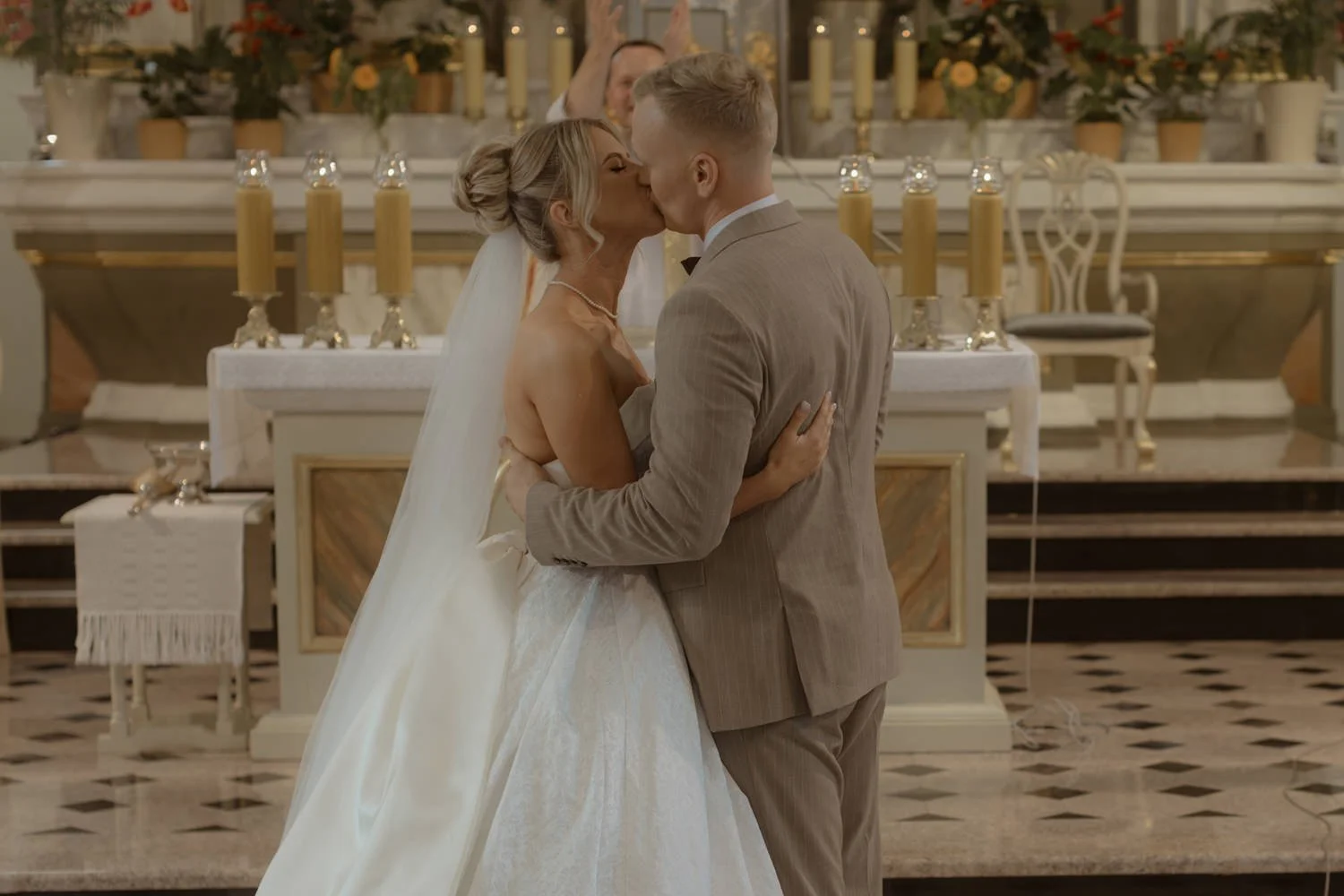 A bride and groom sharing a kiss during their wedding ceremony inside a church, with candles and floral arrangements in the background.