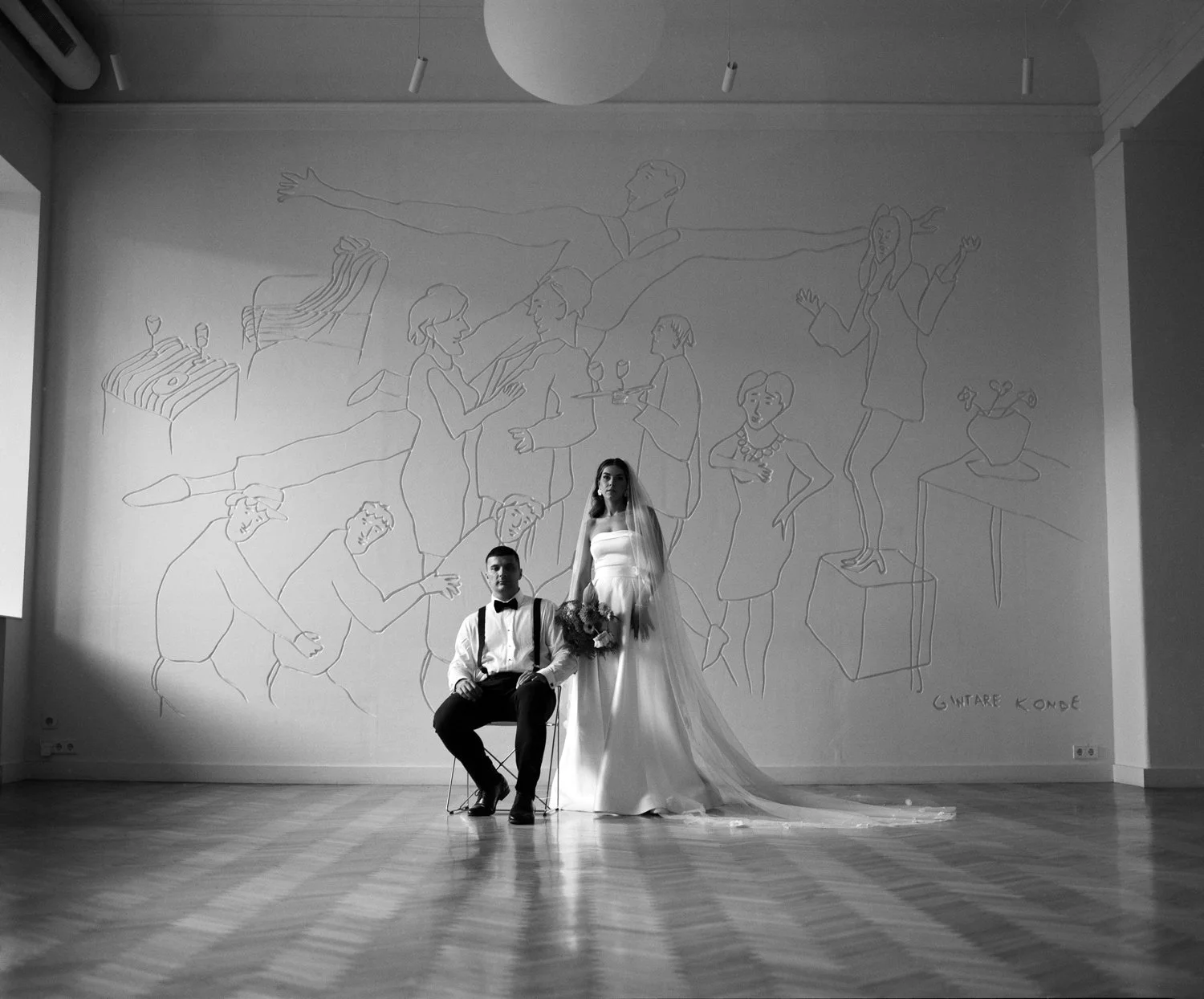 Black and white photo of a bride standing next to a seated groom in front of a large wall with artistic line drawings of people celebrating. The bride is holding a bouquet and wearing a wedding dress with a long train. The groom is wearing a shirt wi
