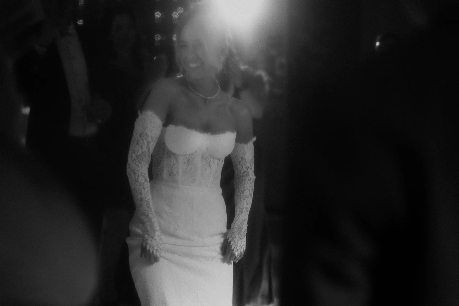 A woman in an elegant white lace dress with long lace sleeves, standing outdoors at night with people in the background, smiling.