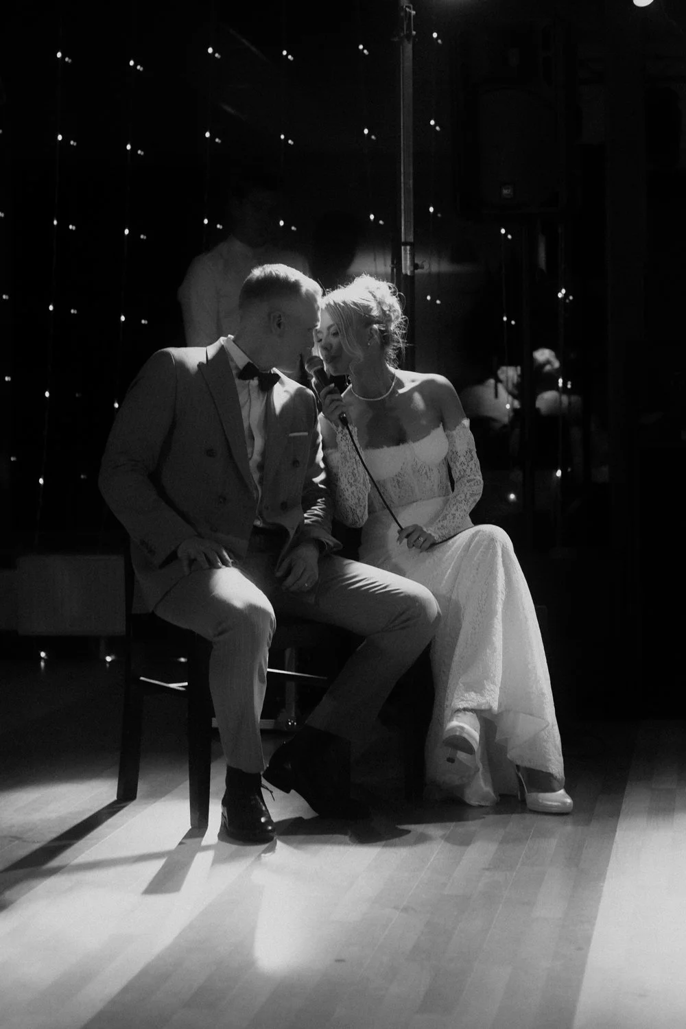 A black and white photo of a bride and groom singing together at their wedding reception, with the bride holding a microphone.