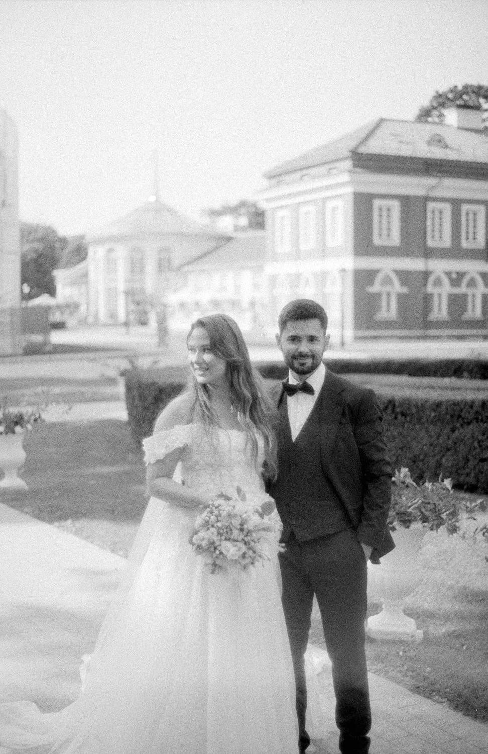 Black and white photo of a bride and groom standing outdoors, with the bride holding a bouquet and the groom in a tuxedo, in front of historic buildings.