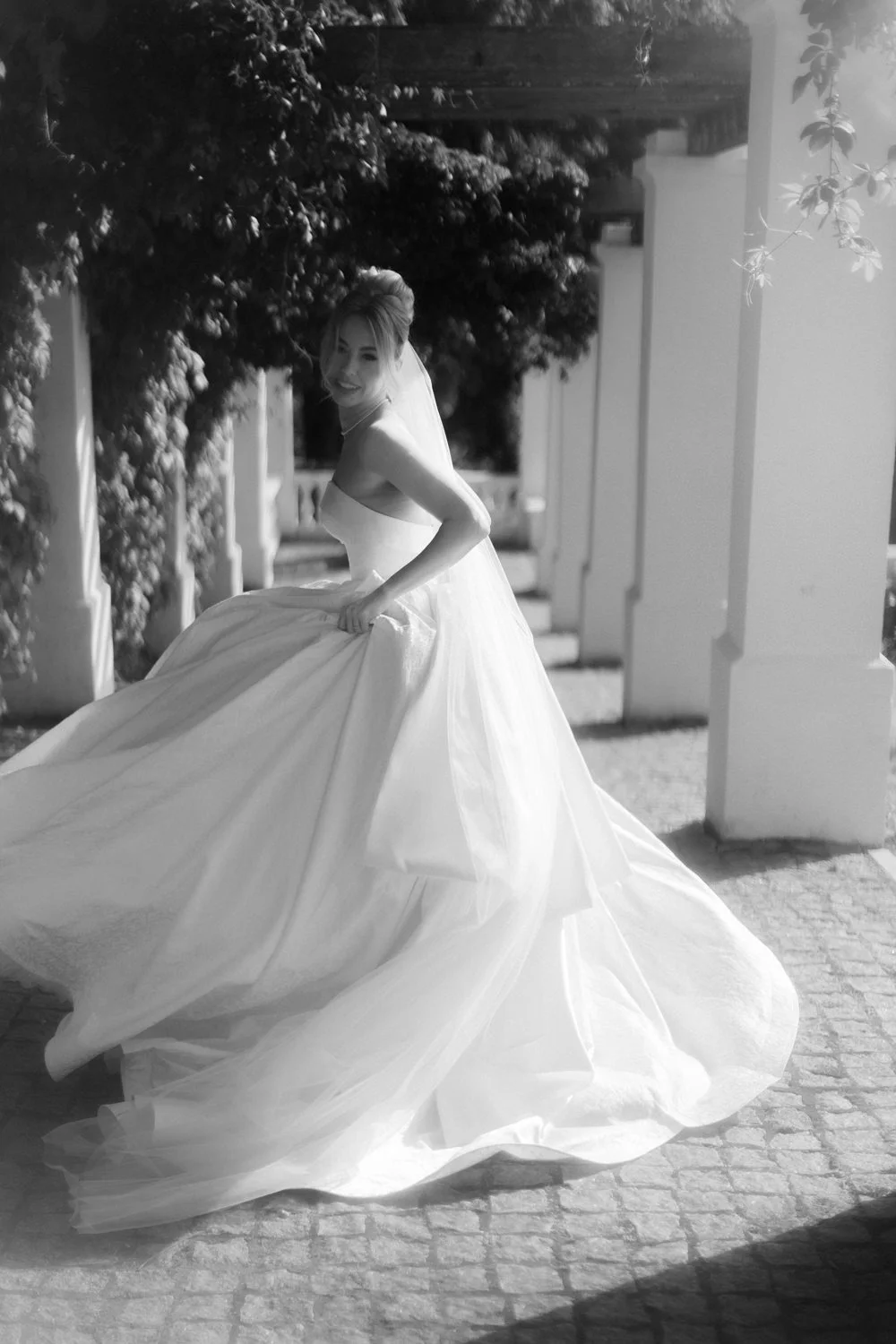 A bride in a wedding dress outdoors, smiling and holding the skirt of her dress.