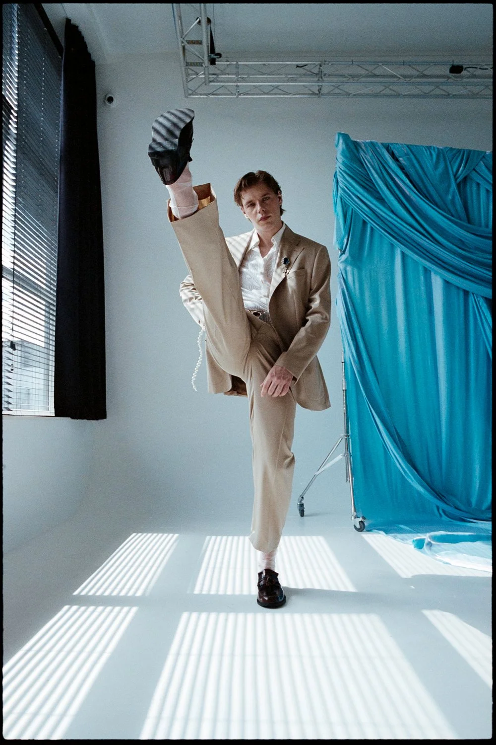 A person in beige suit performing a high kick in front of a blue draped backdrop, with sunlight creating striped shadows on the floor.