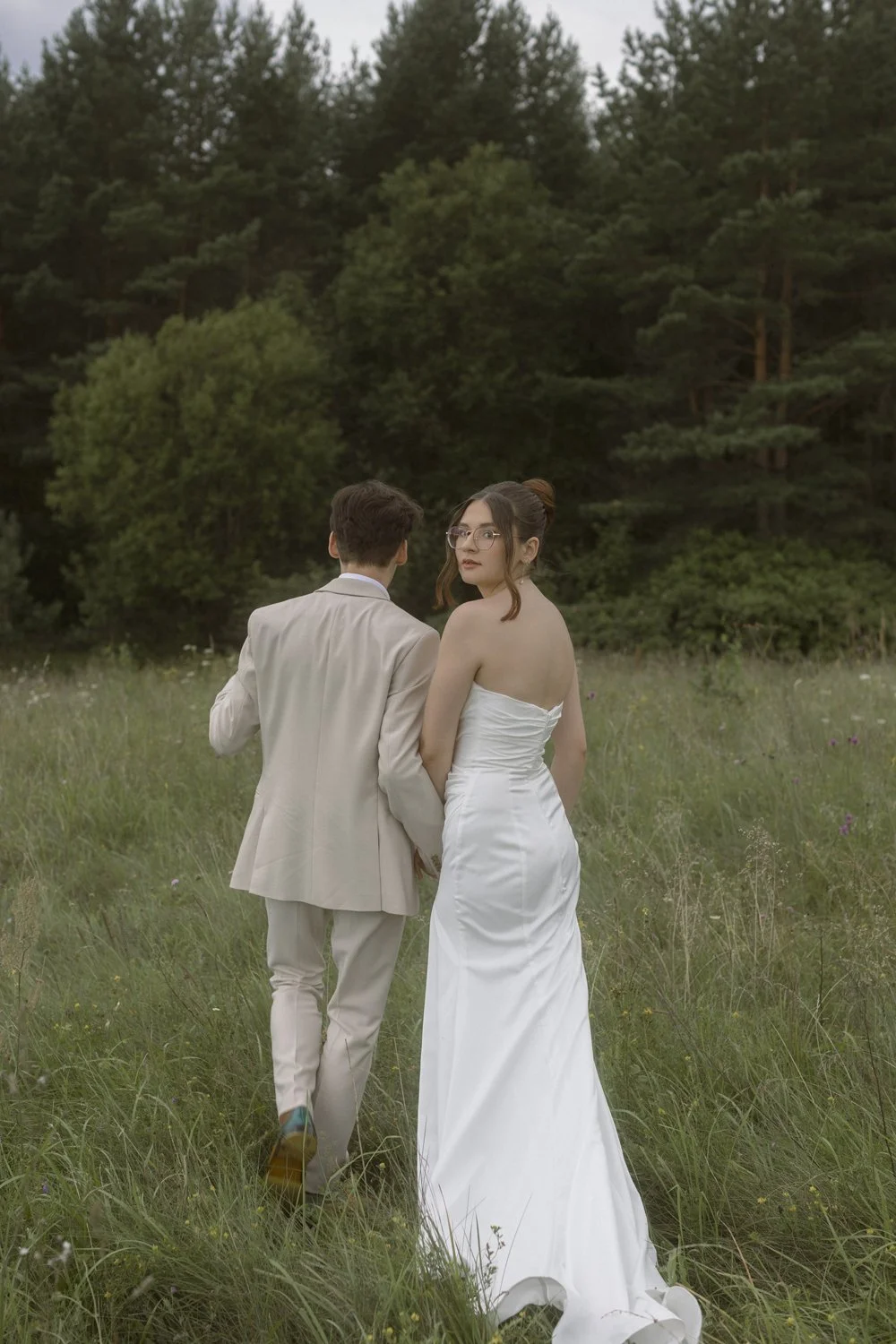 A couple in wedding attire walking through a grassy field with trees in the background. The woman wears a strapless white wedding gown and glasses, while the man is dressed in a beige suit.