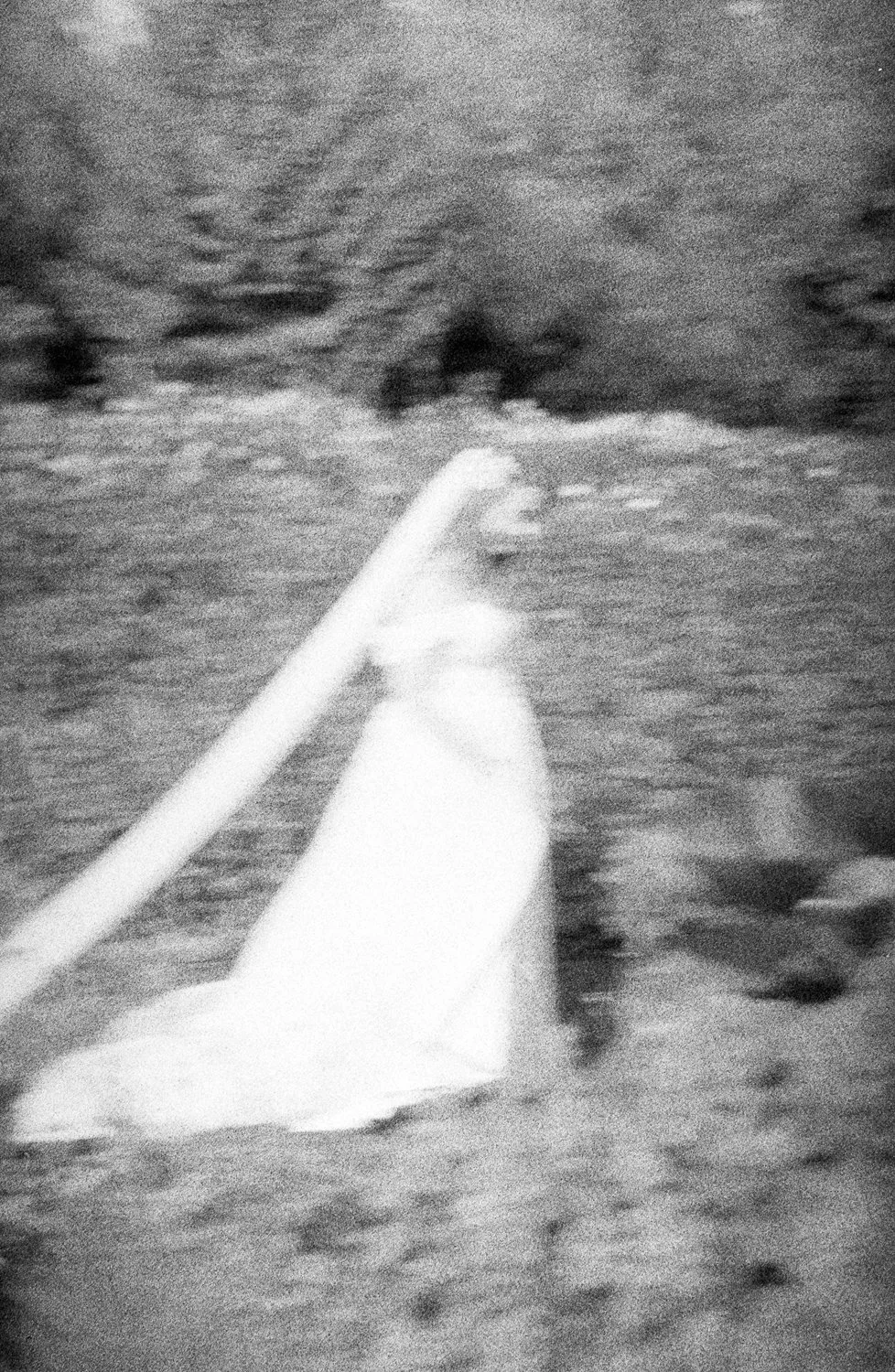 A person in a white dress is lying on the ground on a beach or sandy area, with a white chair beside them. The background shows nature, possibly bushes or trees, with an overall blurred, black-and-white effect.