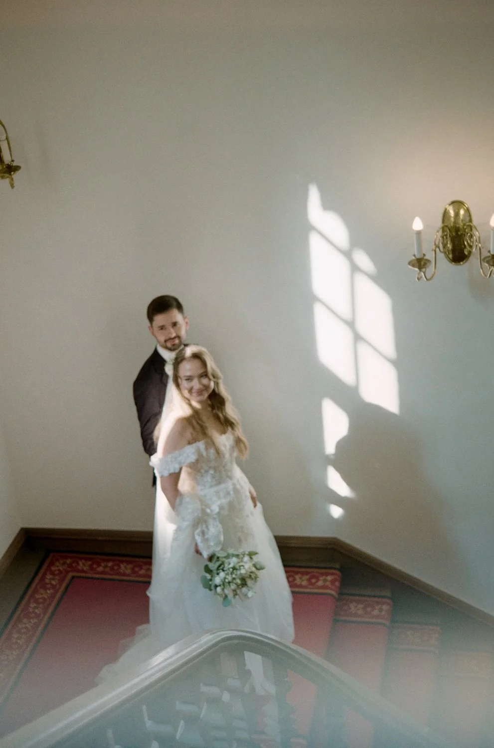 A newlywed couple standing on a staircase. The bride is wearing a white wedding gown with lace details, holding a bouquet of white flowers, and smiling. The groom is wearing a dark suit and smiling as well. Sunlight coming through a window creates a 