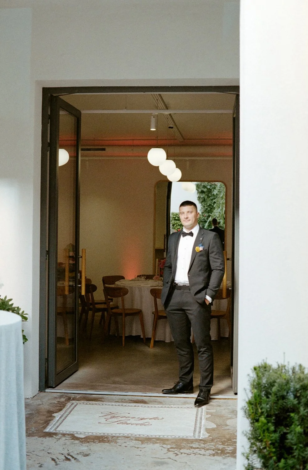 A young man in a tuxedo standing in the doorway of a decorated event room, with a round table and white tablecloth visible inside.