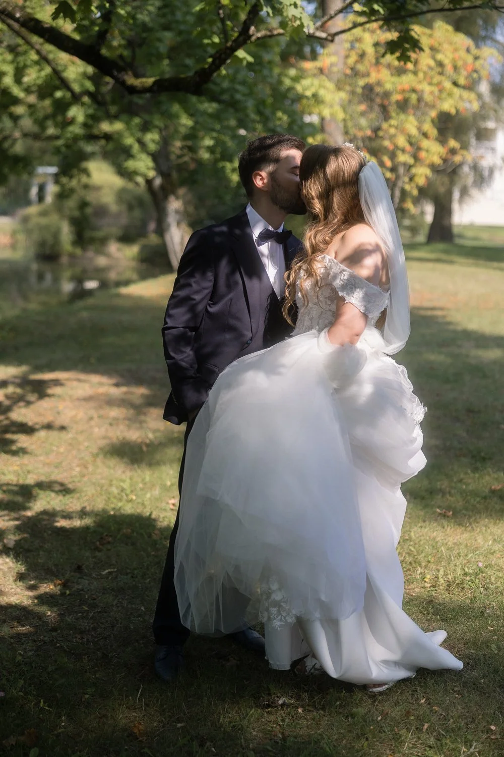 A bride and groom sharing a kiss outdoors in a park setting, with trees and sunlight in the background.