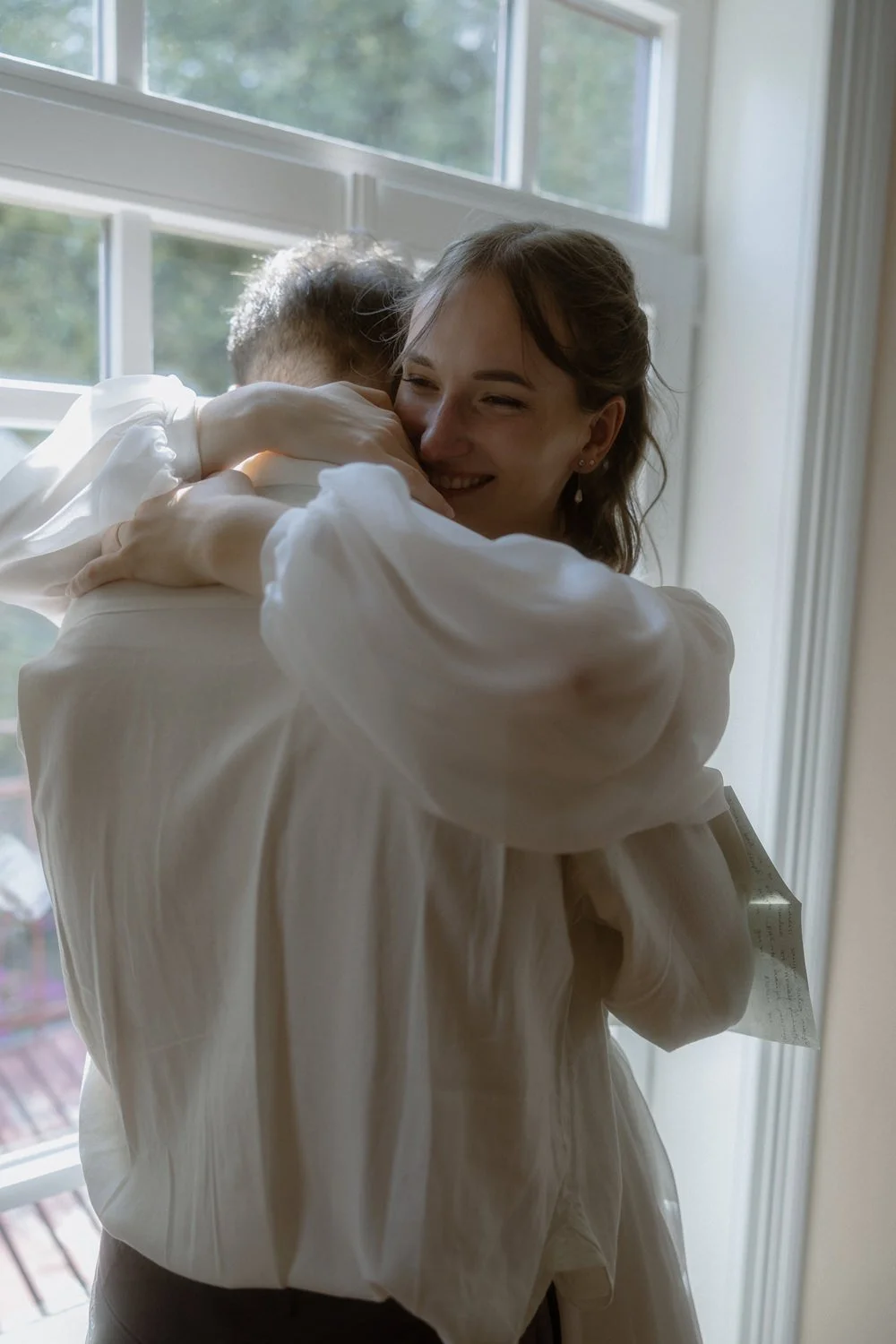 A woman is hugging a small child near a window, smiling warmly, with natural daylight illuminating the scene.