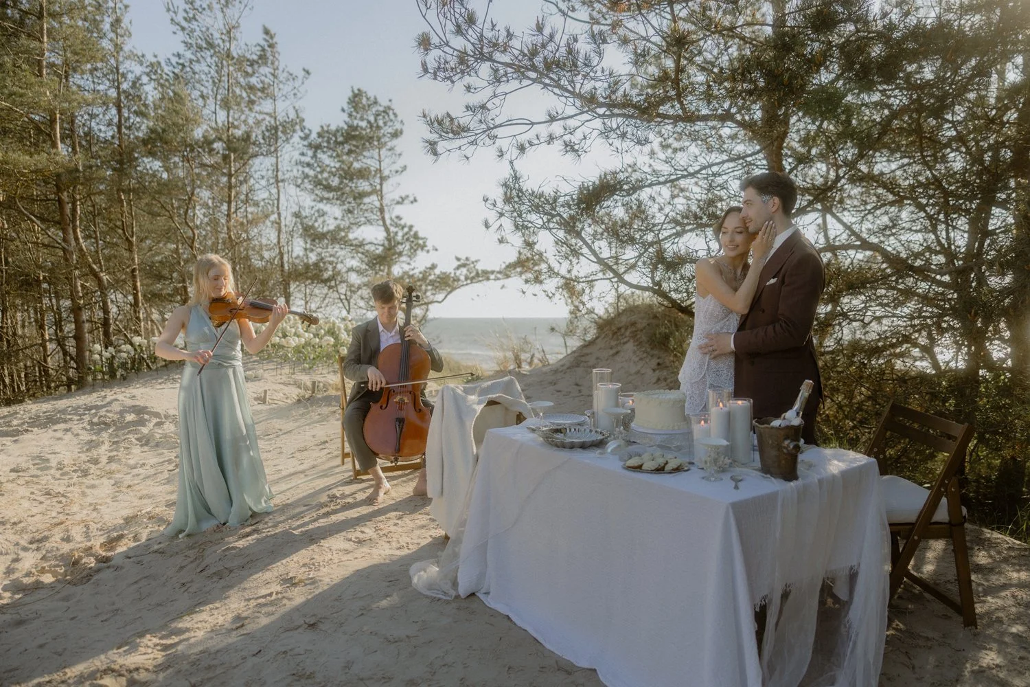 A couple at their wedding reception on a sandy beach, with musicians playing violin and cello in the background, a table with a cake, candles, and snacks, surrounded by trees and overlooking the ocean during sunset.