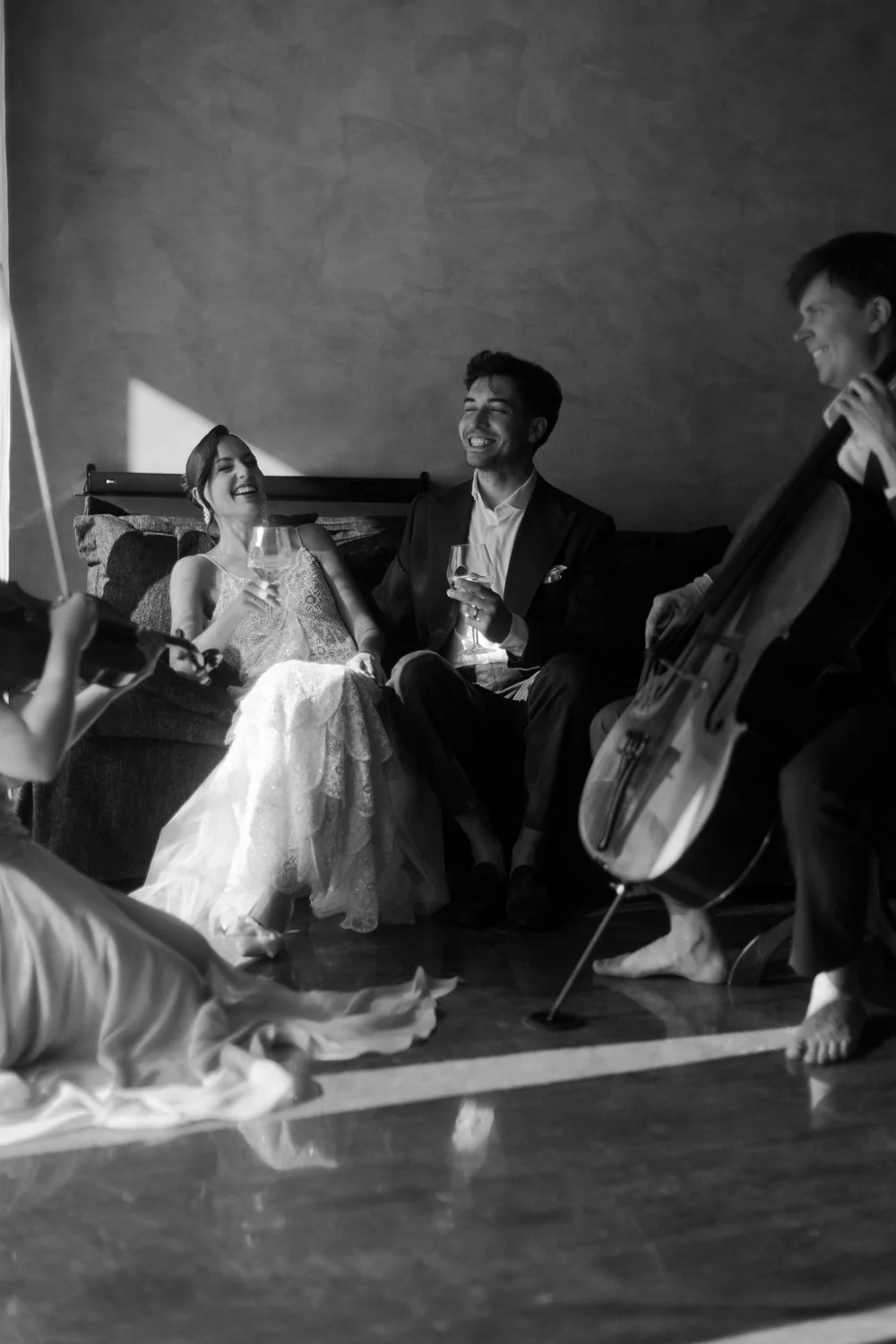 A wedding reception with a bride and groom sitting on a couch, smiling and holding wine glasses, while a cellist plays nearby, and a person in the foreground writes on a notepad.