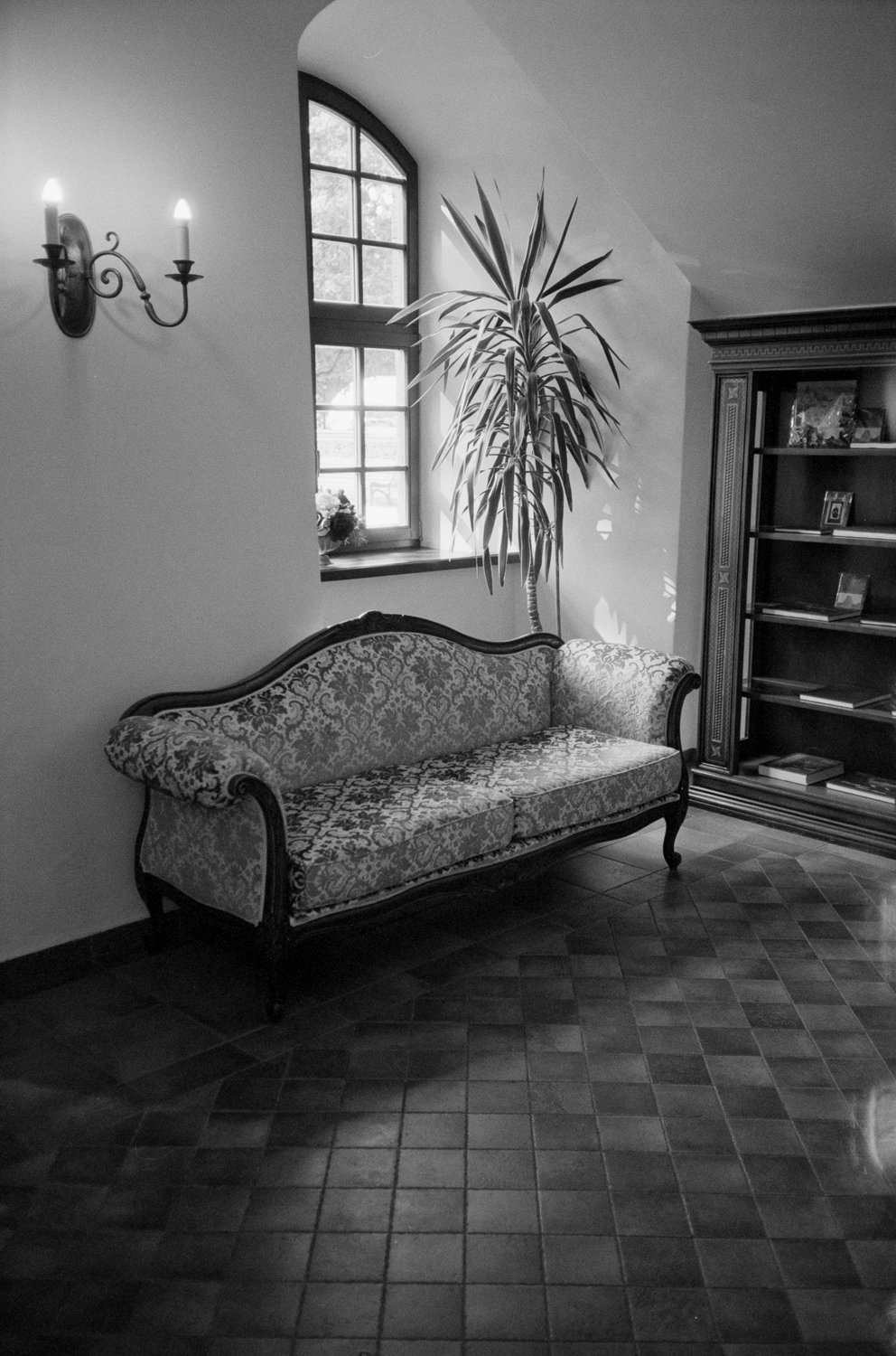 An interior room with a vintage floral sofa, a window with a large plant nearby, a wooden bookcase, a wall sconce with two candles, and a tiled floor.