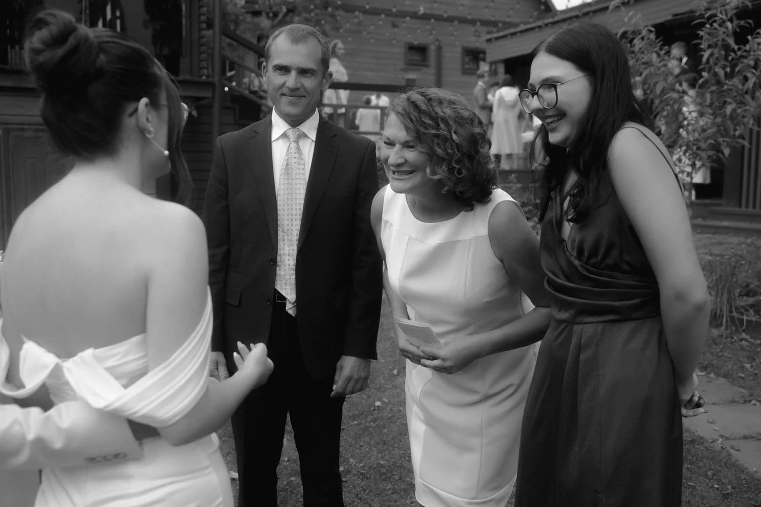 Four adults smiling and engaging in conversation outdoors at a social event, with three women and one man, some dressed formally, in a garden setting.