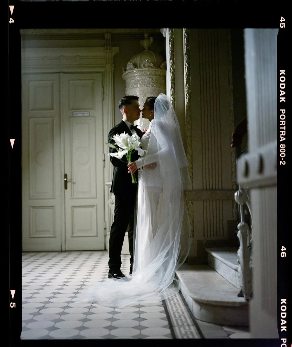 A bride and groom standing close together indoors, facing each other. The bride is dressed in a white wedding gown with a veil and holding a bouquet of white lilies. The groom is wearing a black tuxedo. They are standing in front of ornate architecture with a checkered floor.