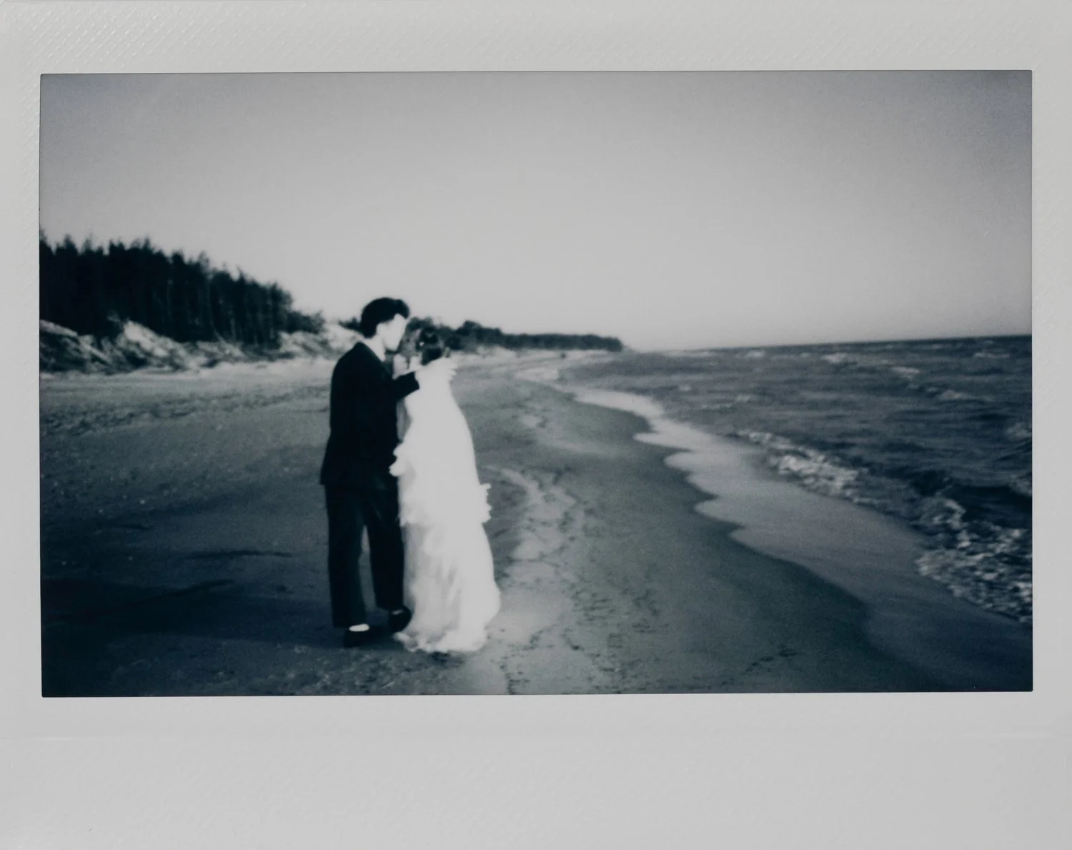 A black and white photo of a couple in formal wedding attire standing on the beach near the shoreline, with the ocean waves approaching them and trees in the background.