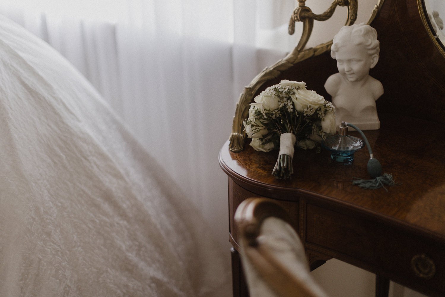 A vintage wooden bedside table with decorative gold accents, holding a small bouquet of white roses, a sculpted bust of a child's head, and a blue perfume bottle with a spray nozzle in an elegantly decorated room.