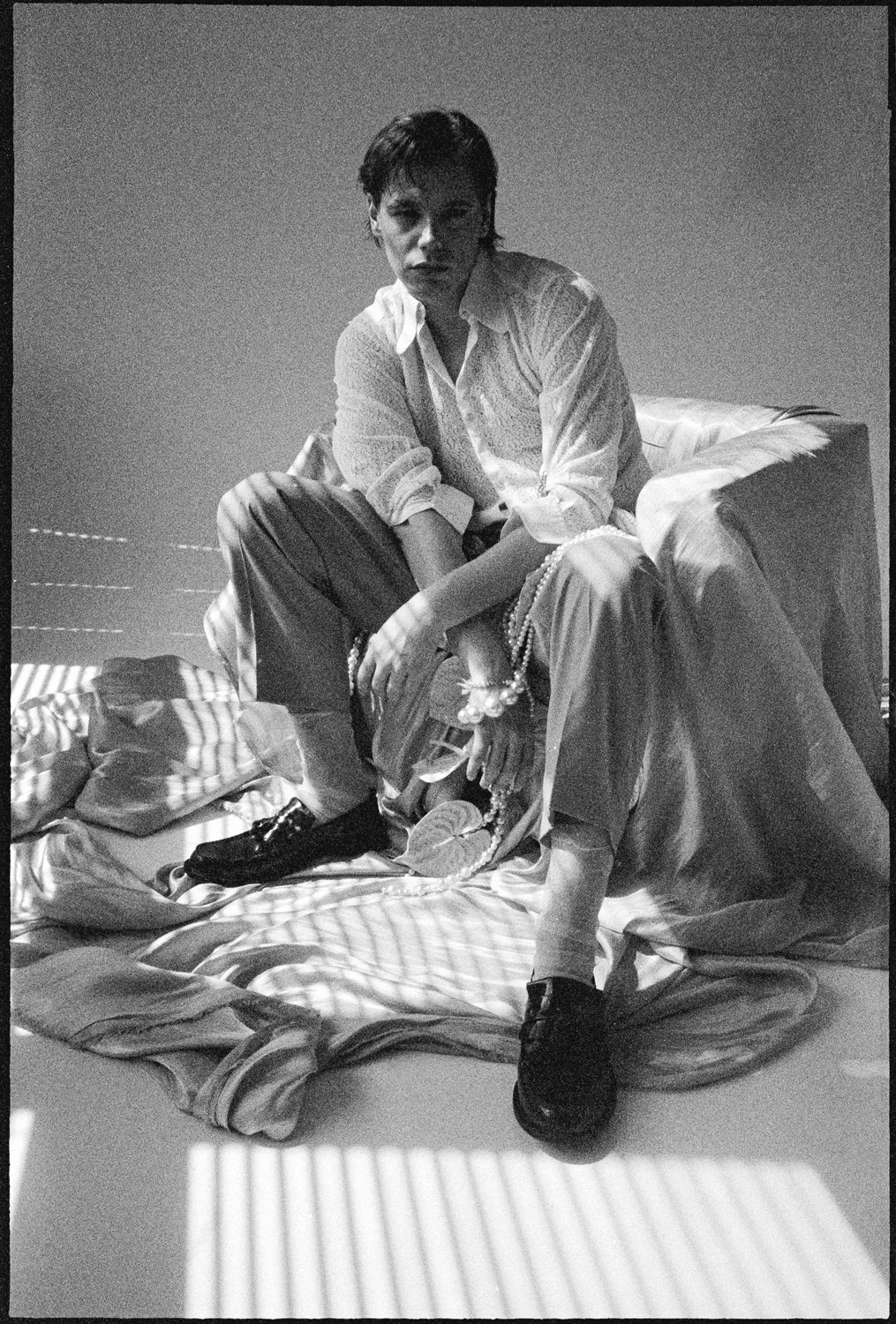 A black and white photo of a person sitting on a bed with scattered clothes. They have short hair and are wearing a button-up shirt and loose pants, looking directly at the camera.