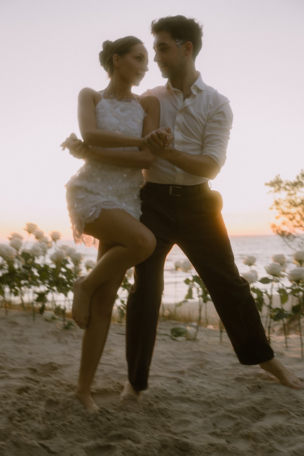 A couple dancing on the beach at sunset, with the ocean and flowers in the background.