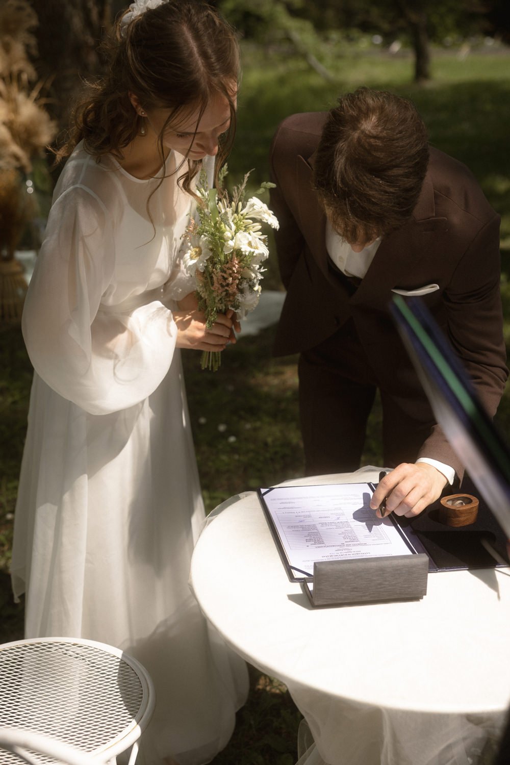 A bride in a white dress holding a bouquet of flowers while a man in a brown suit signs documents at a small outdoor wedding ceremony.