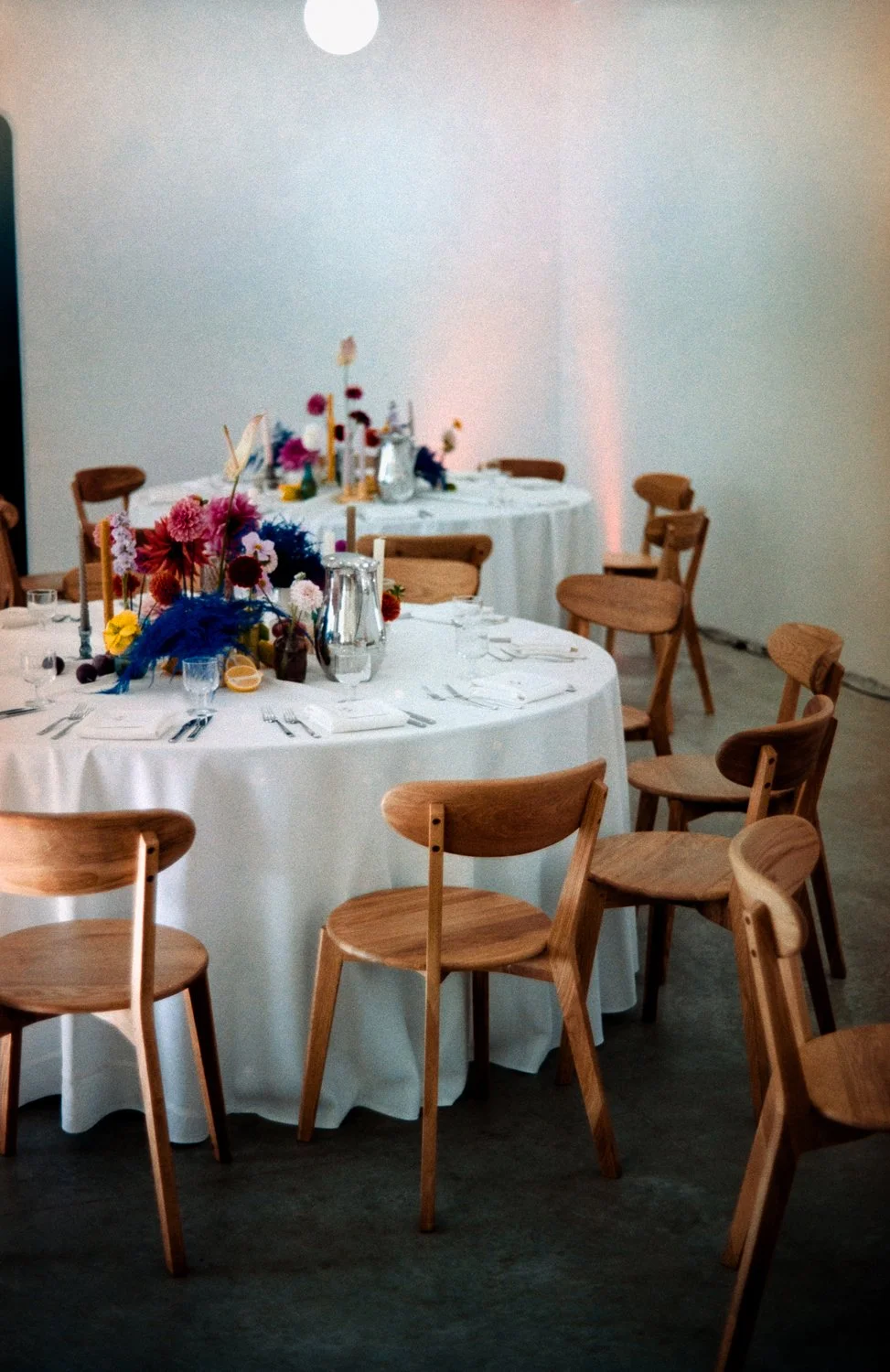 Indoor event space with round tables dressed in white tablecloths, decorated with colorful floral centerpieces, surrounded by wooden chairs.